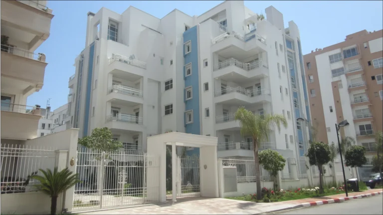 Modern apartment building in Casablanca with white and blue facade, multiple balconies, gated entrance, and palm trees