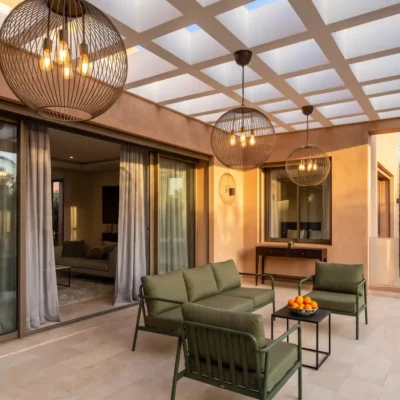 Villa covered terrace at dusk with green outdoor sofas, spherical pendant lights and garden view beyond glass rail